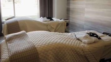 A hotel room with two beds, each with a quilted bedspread and pillows. There is a window with curtains, a wall-mounted shelf with toiletries, and a view of the outdoors.