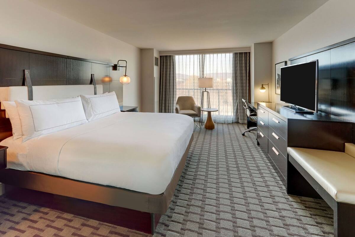 A hotel room with a large bed, a desk with a TV, a chair, and a view of the city through the window.