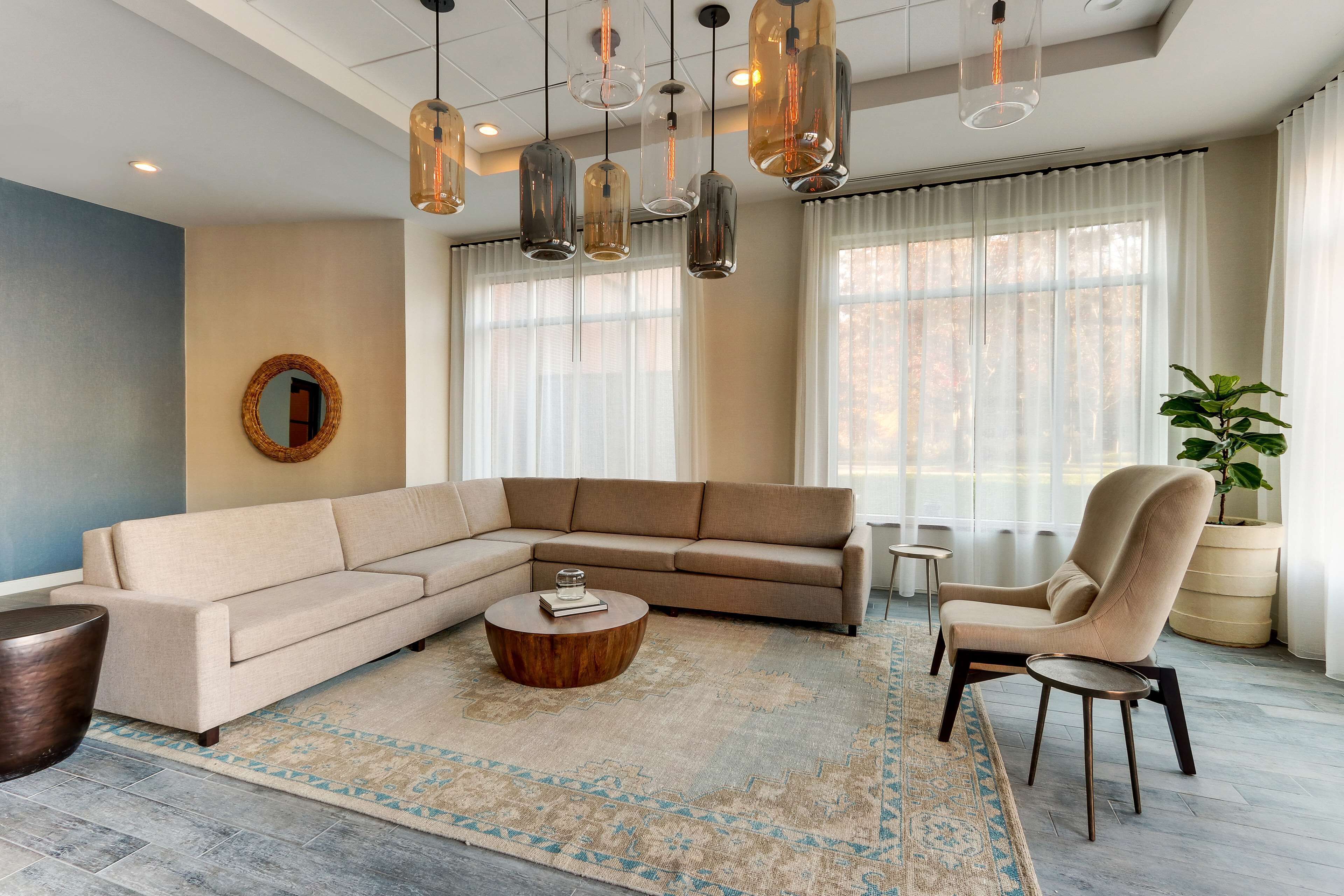 Een moderne woonkamer met een hoekbank, een rond bijzettafeltje en een stoel. Er hangen pendelverlichting, een spiegel aan de muur, en een groot raam met dunne gordijnen.