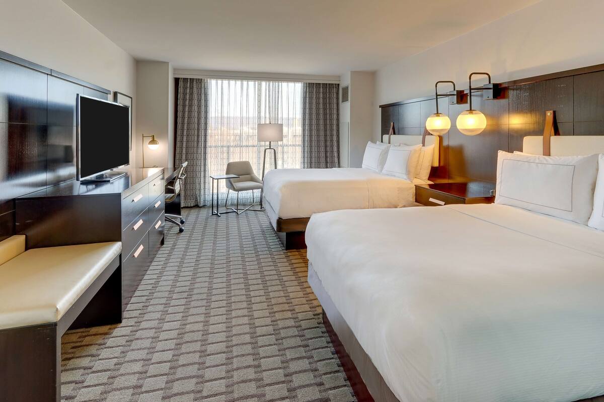A hotel room with a large bed, a smaller bed, a desk with a TV, and a chair.