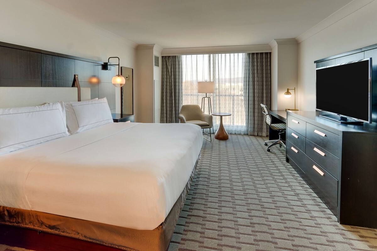 A hotel room with a large bed, a desk with a chair, a television, and a window with curtains.