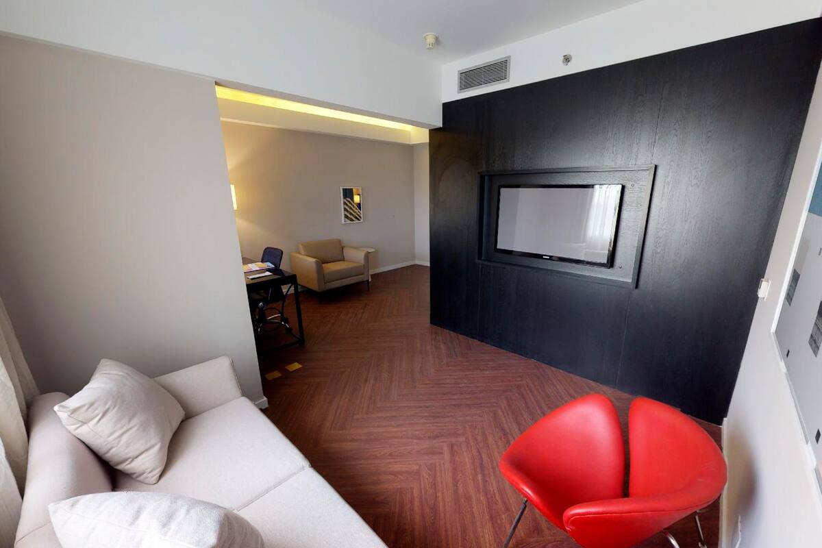 Quarto de hotel moderno com sofá, uma cadeira vermelha, televisão presa a uma parede escura e chão de madeira.