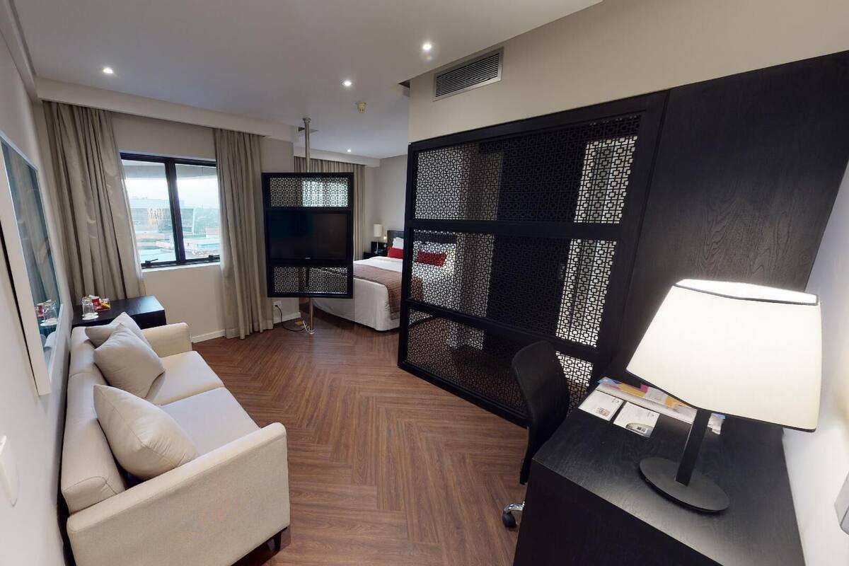 Quarto de hotel com sofá, escrivaninha com luminária, televisão e cama com vista para a cidade.