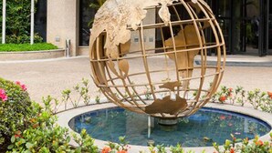 Una escultura de un globo terráqueo en una fuente, rodeada de follaje, frente a un edificio con un letrero que dice "ESTANPLAF FUNCHAL-FARIAL".