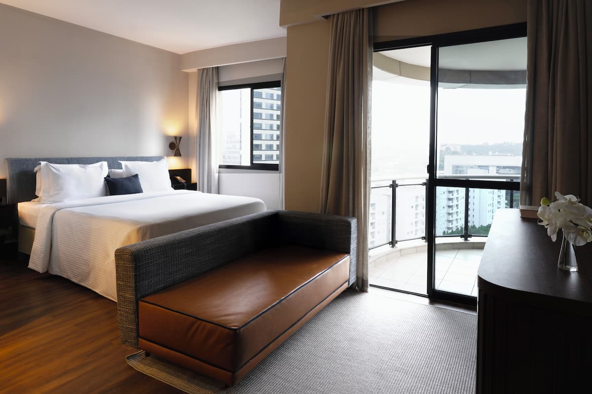 Quarto de hotel com cama grande, sofá, escrivaninha e varanda com vista para a cidade.