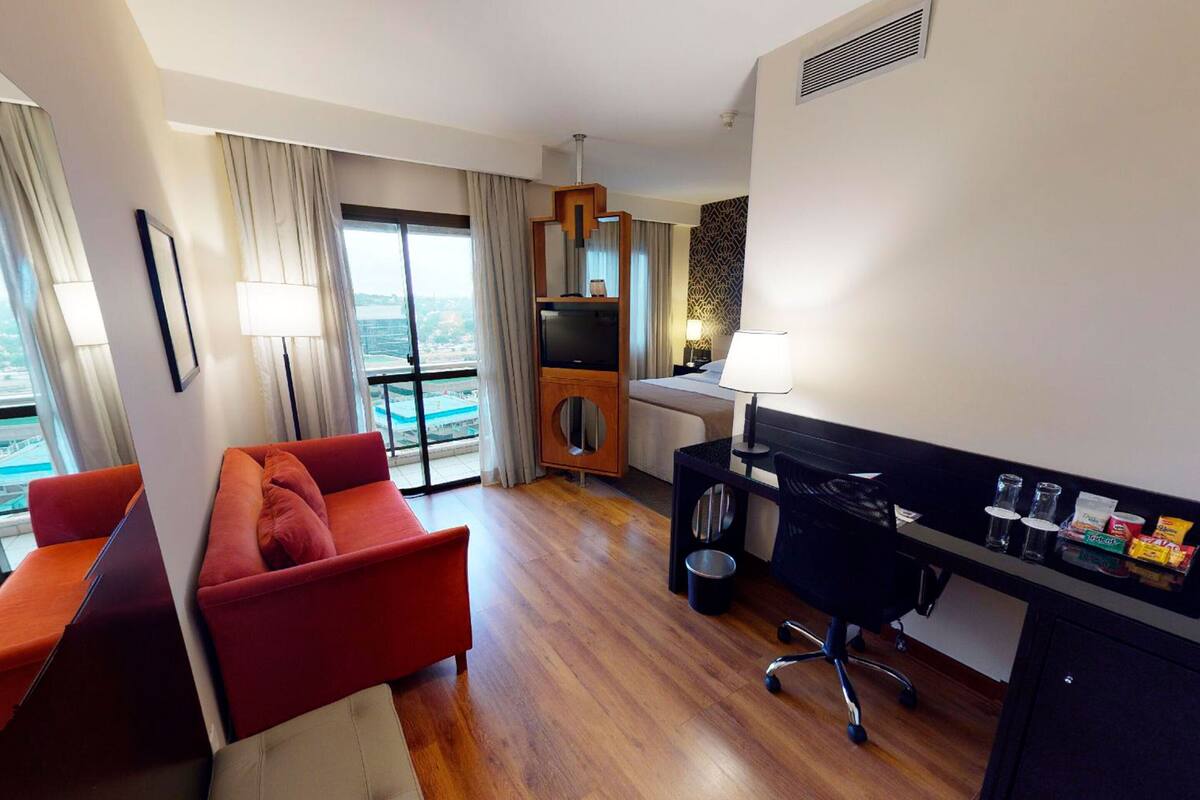 Quarto de hotel moderno com um sofá vermelho, uma escrivaninha com computador, uma televisão e uma varanda com vista.