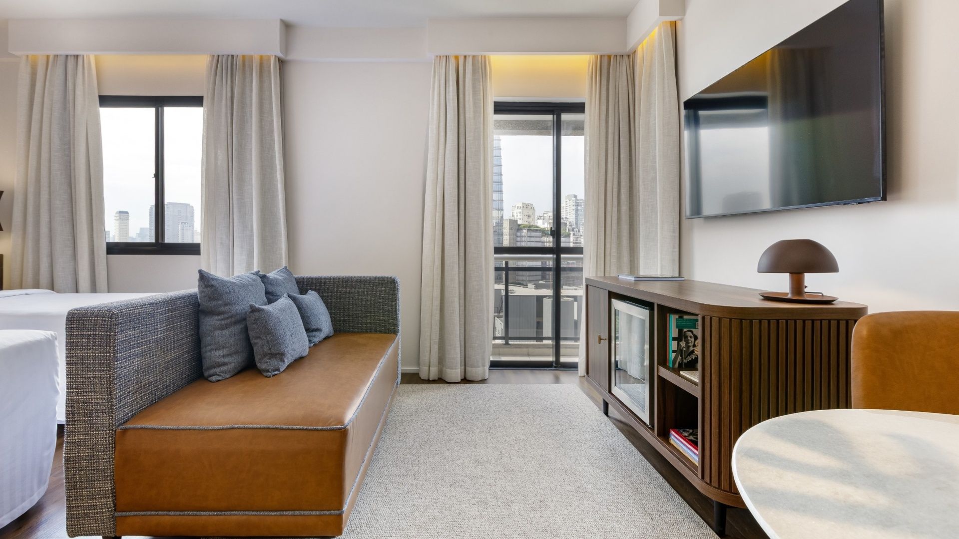 A hotel room with a sofa, a television, a cabinet, and a view of the city.