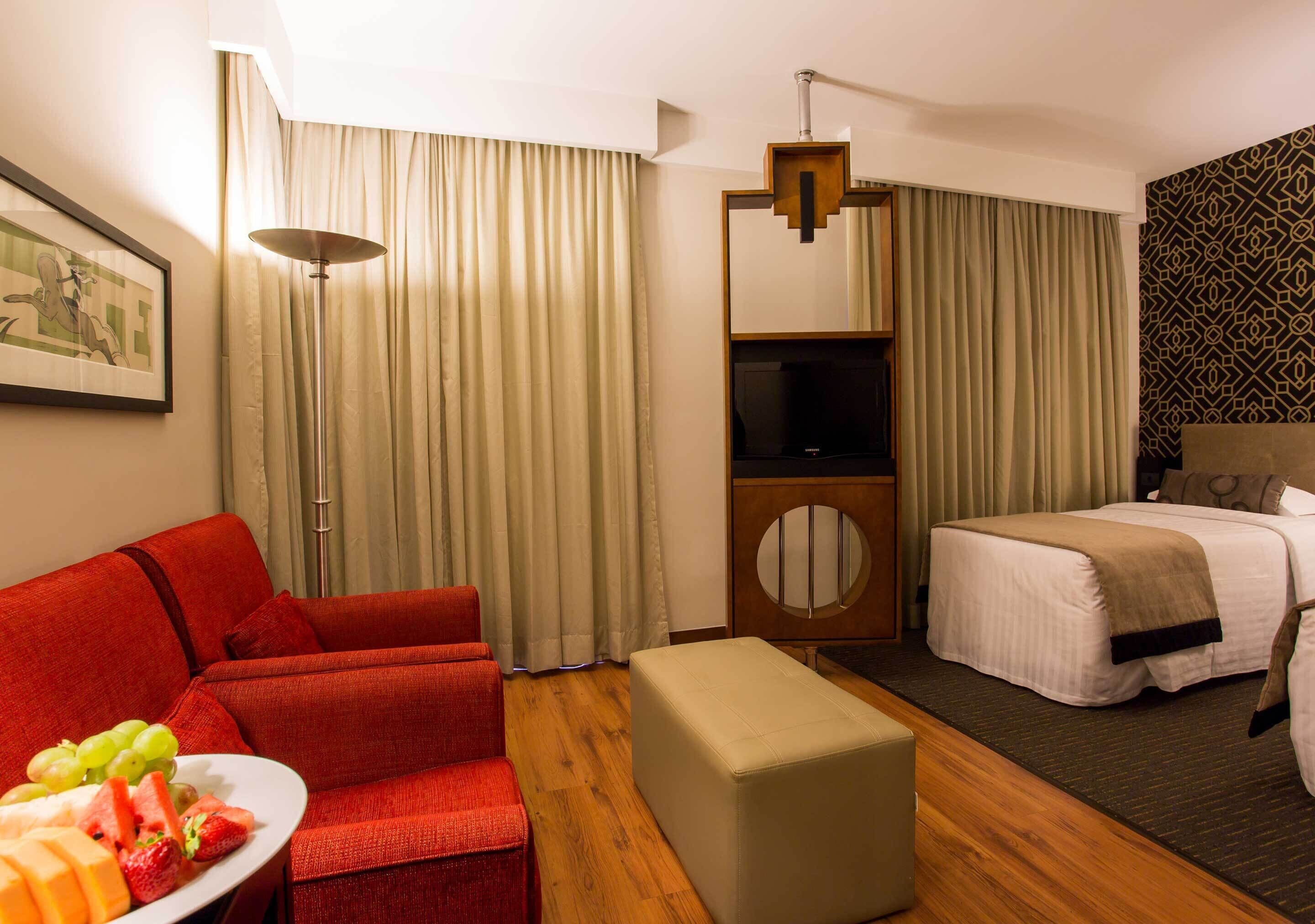 A hotel room with a red sofa, a bed, a TV, and a dining table with fruit.