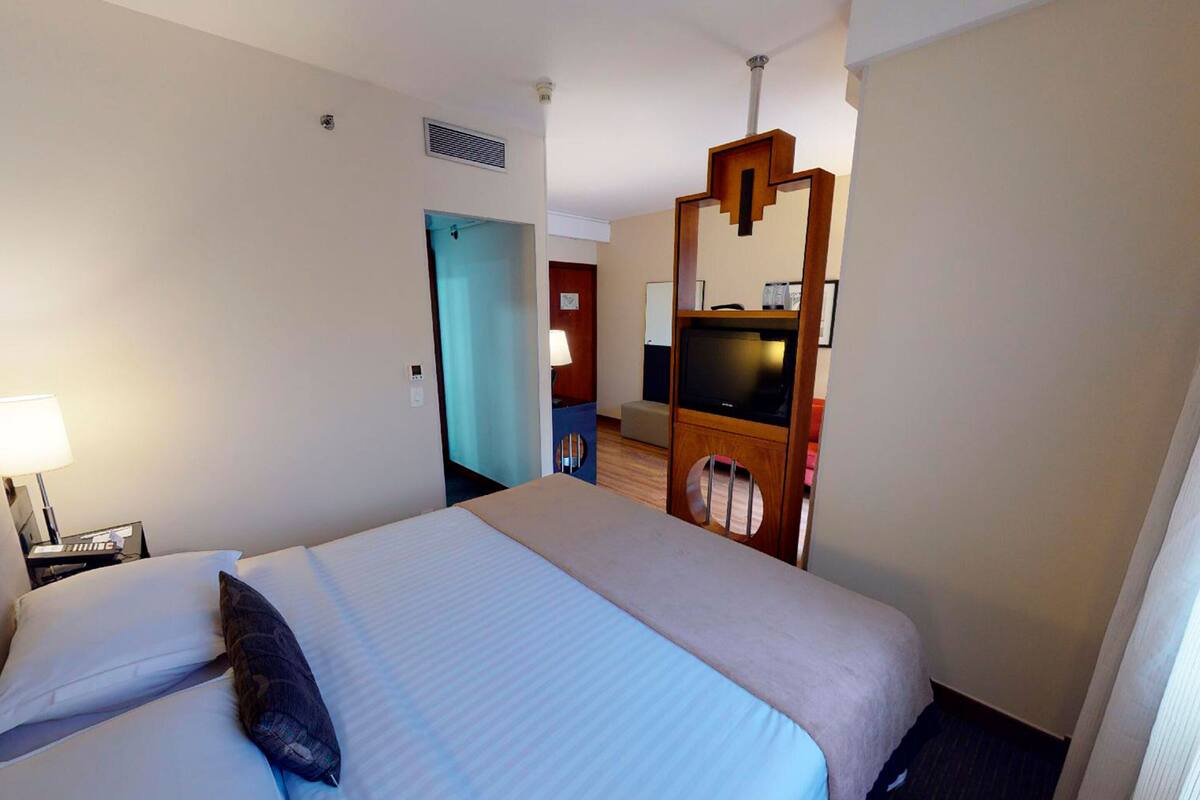 Quarto de hotel com cama, televisão, secretária com abajur e porta para outro espaço.