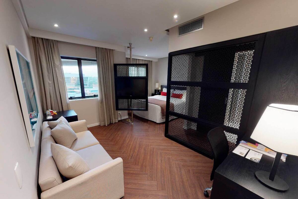 Quarto de hotel moderno com sofá, escrivaninha com luminária, TV e uma grande janela com vista.