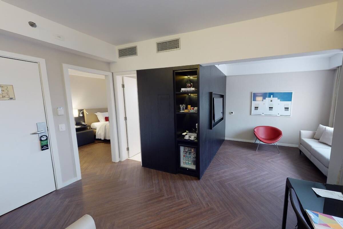 Quarto de hotel com chão de madeira, um móvel de armazenamento preto, uma cadeira vermelha, um sofá branco e um quadro pendurado na parede.