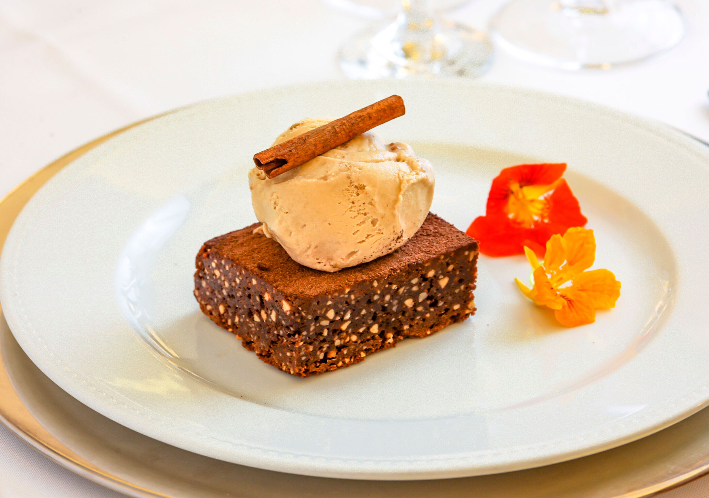 Une assiette à dessert avec une part de gâteau, surmontée d’une boule de glace et d’une branche de cannelle, accompagnée d’une fleur.