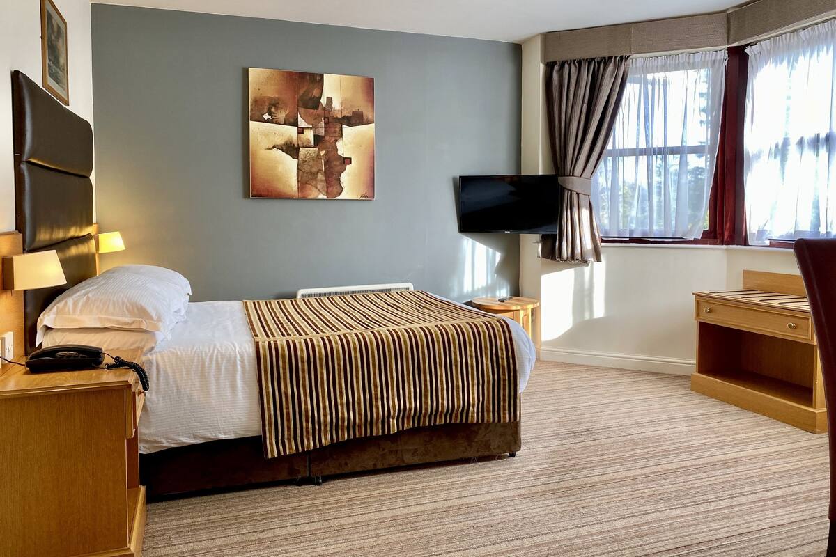 A hotel room with a bed, bedside tables, a TV, a painting on the wall, and a window with curtains.