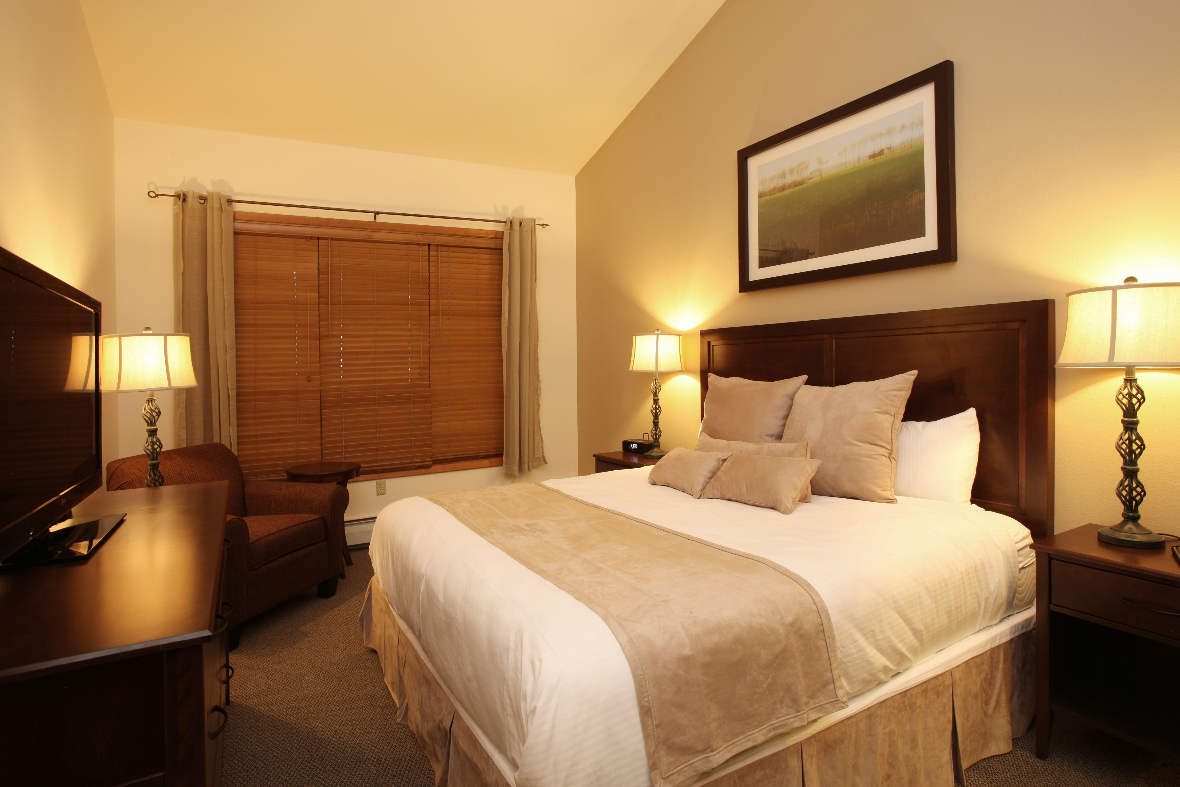 Une chambre d’hôtel avec un grand lit, deux tables de chevet avec des lampes, une tête de lit en bois, un bureau avec une chaise et un téléviseur.