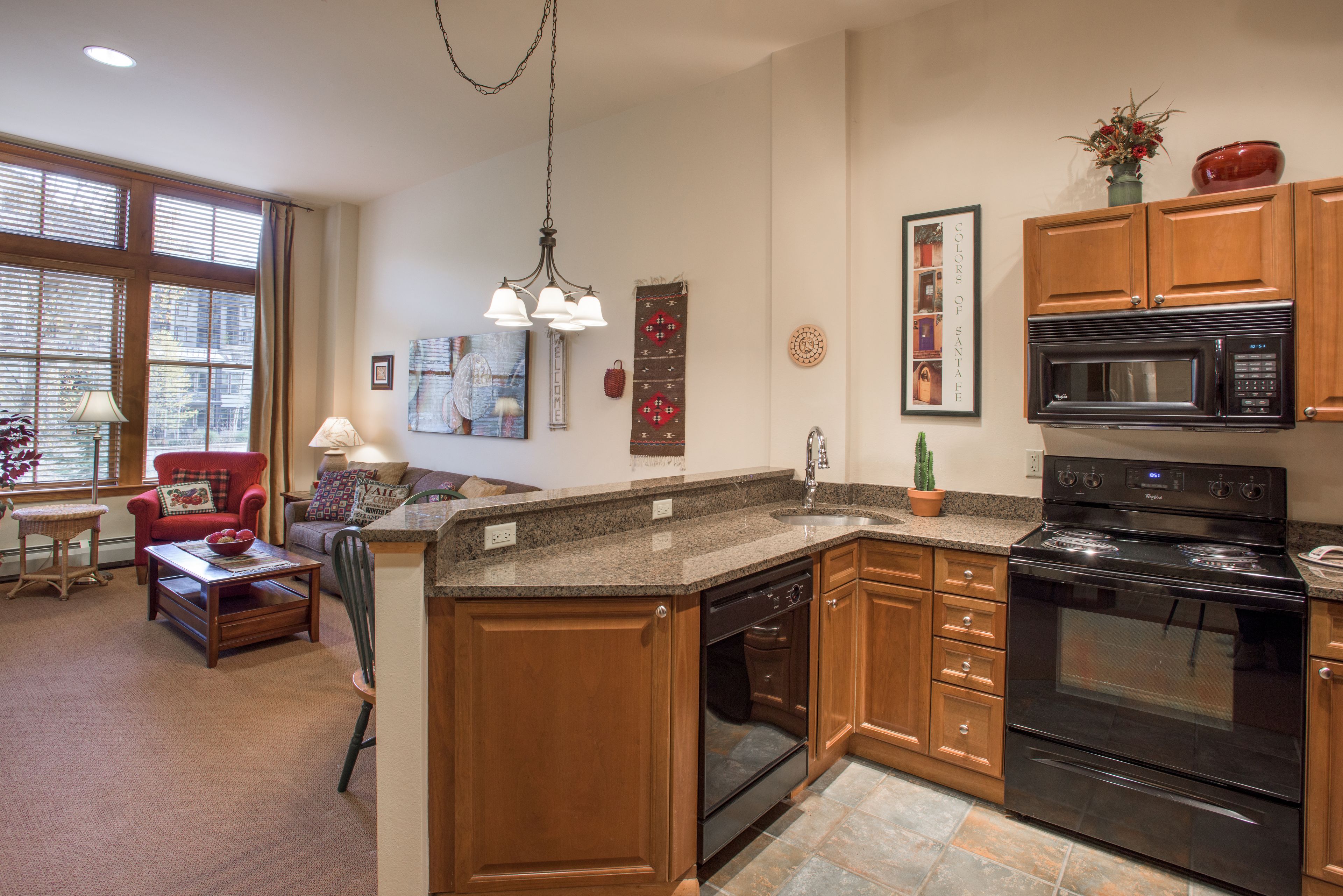 A kitchen with wooden cabinets, a black microwave, and a stove. There is a dining area with a red chair and a lamp.