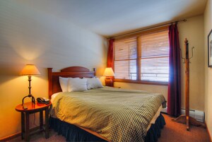 Una cama bien hecha con colcha a cuadros, dos lámparas de noche, una mesita con teléfono y una ventana con persianas y cortinas.