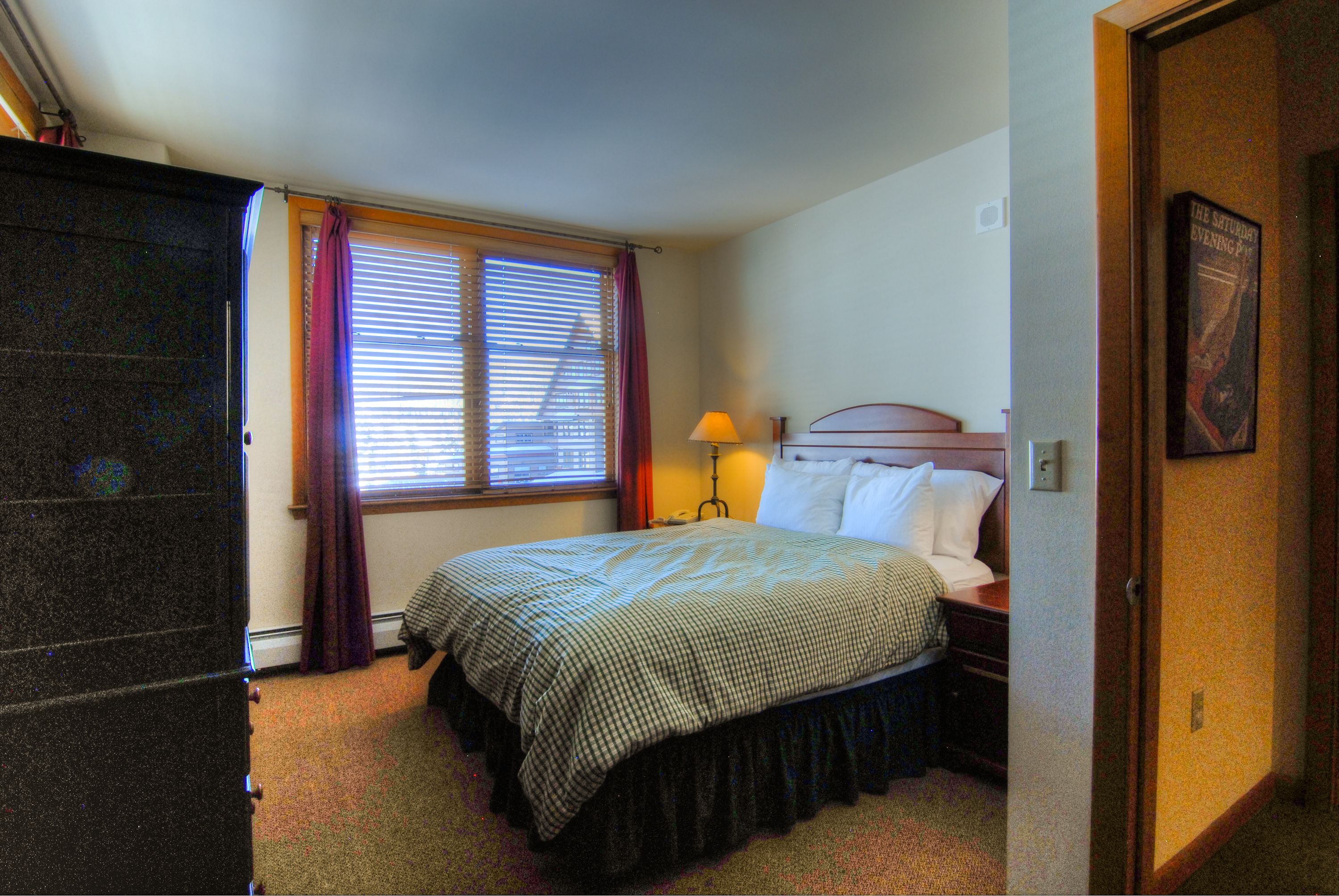 Quarto de hotel com cama, mesa de cabeceira, abajur e janela com persianas.