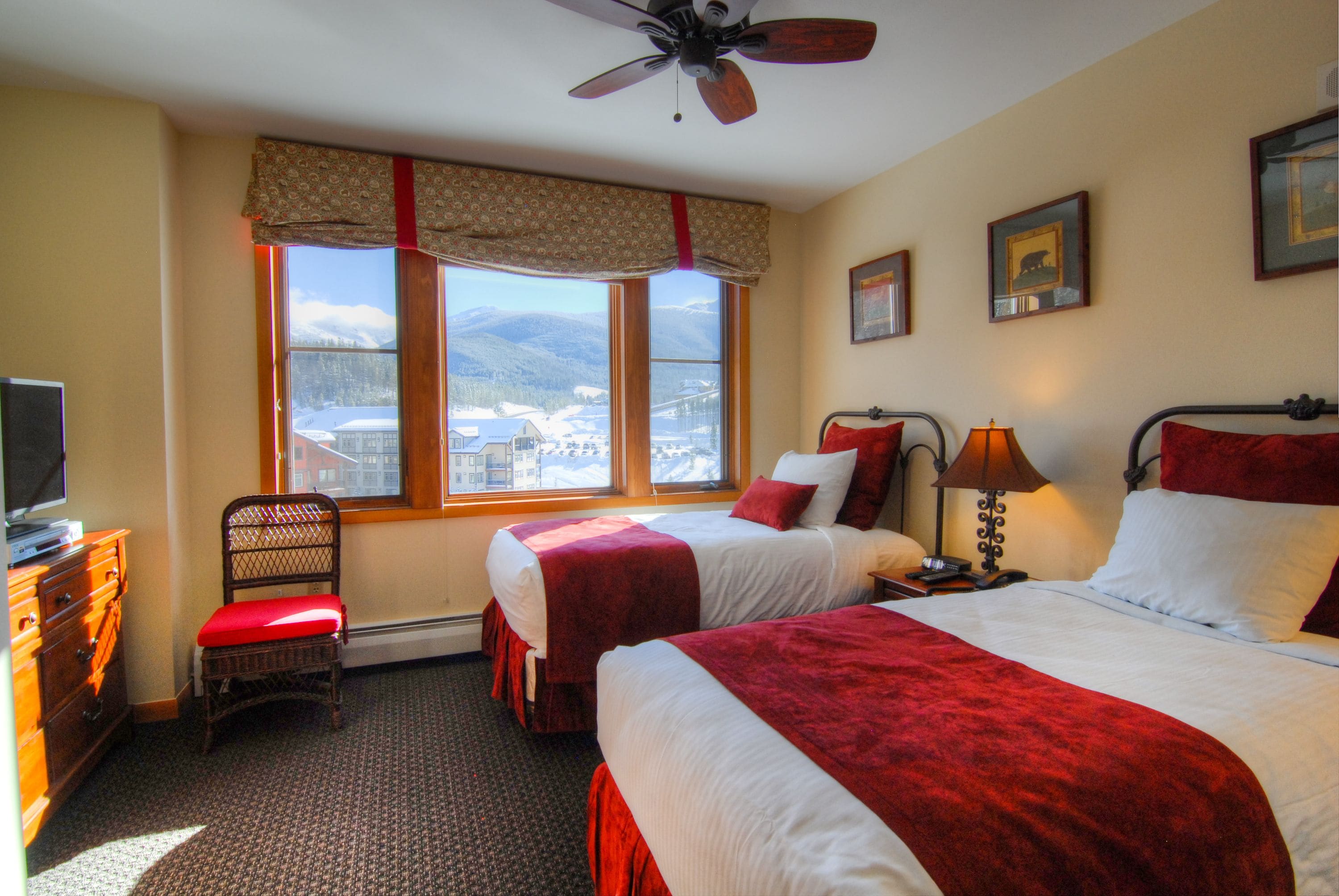 A hotel room with two beds, a chair, a desk with a lamp, a ceiling fan, and a window with a mountain view.