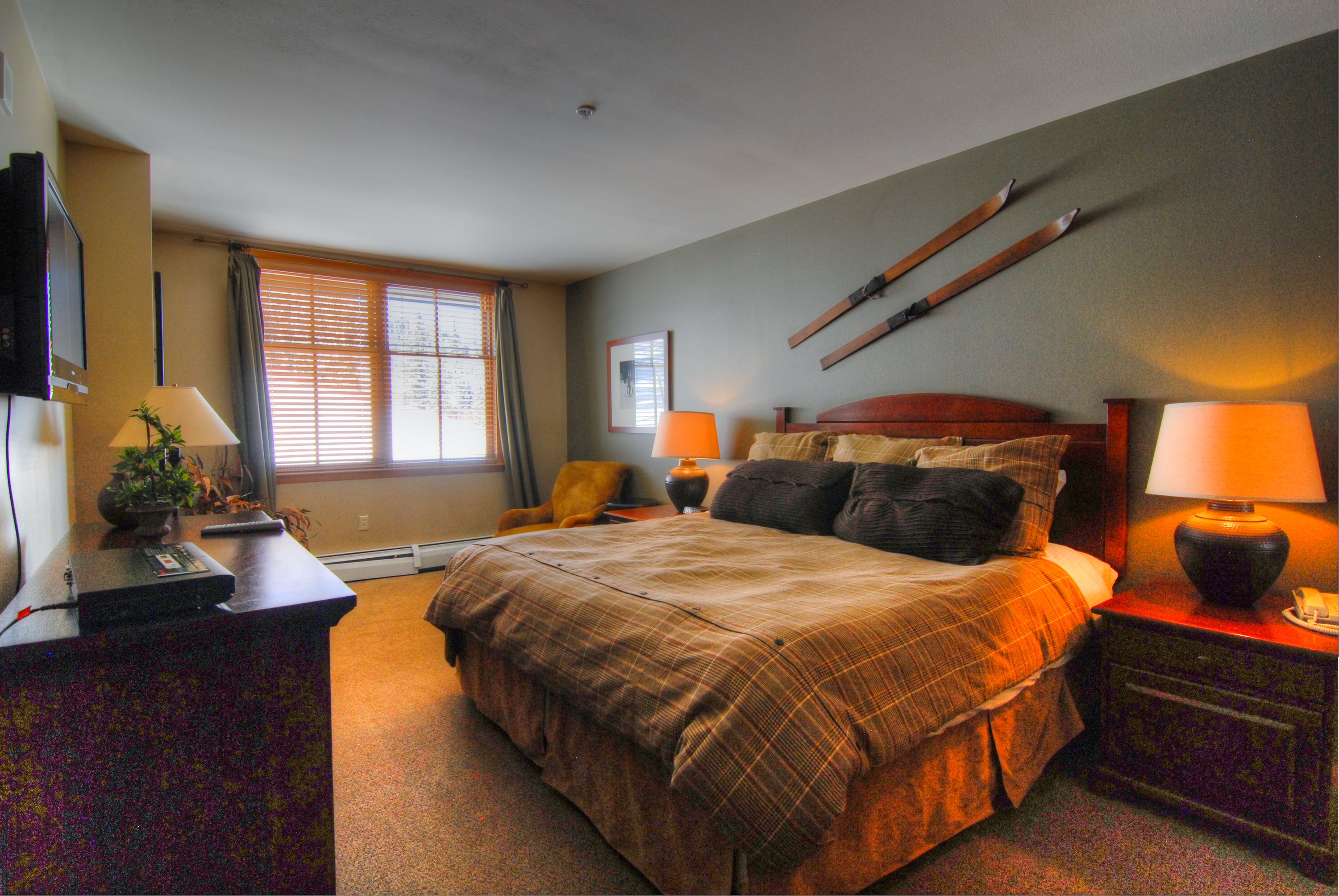 A hotel room with a bed, two bedside lamps, a desk with a computer, a chair, a TV, and skis mounted on the wall.