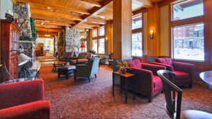 A lobby with wooden beams, stone walls, and large windows. There are red and grey chairs, a fireplace, and a stone pillar with a Christmas tree. - Zephyr Mountain Lodge (Winter Park)
