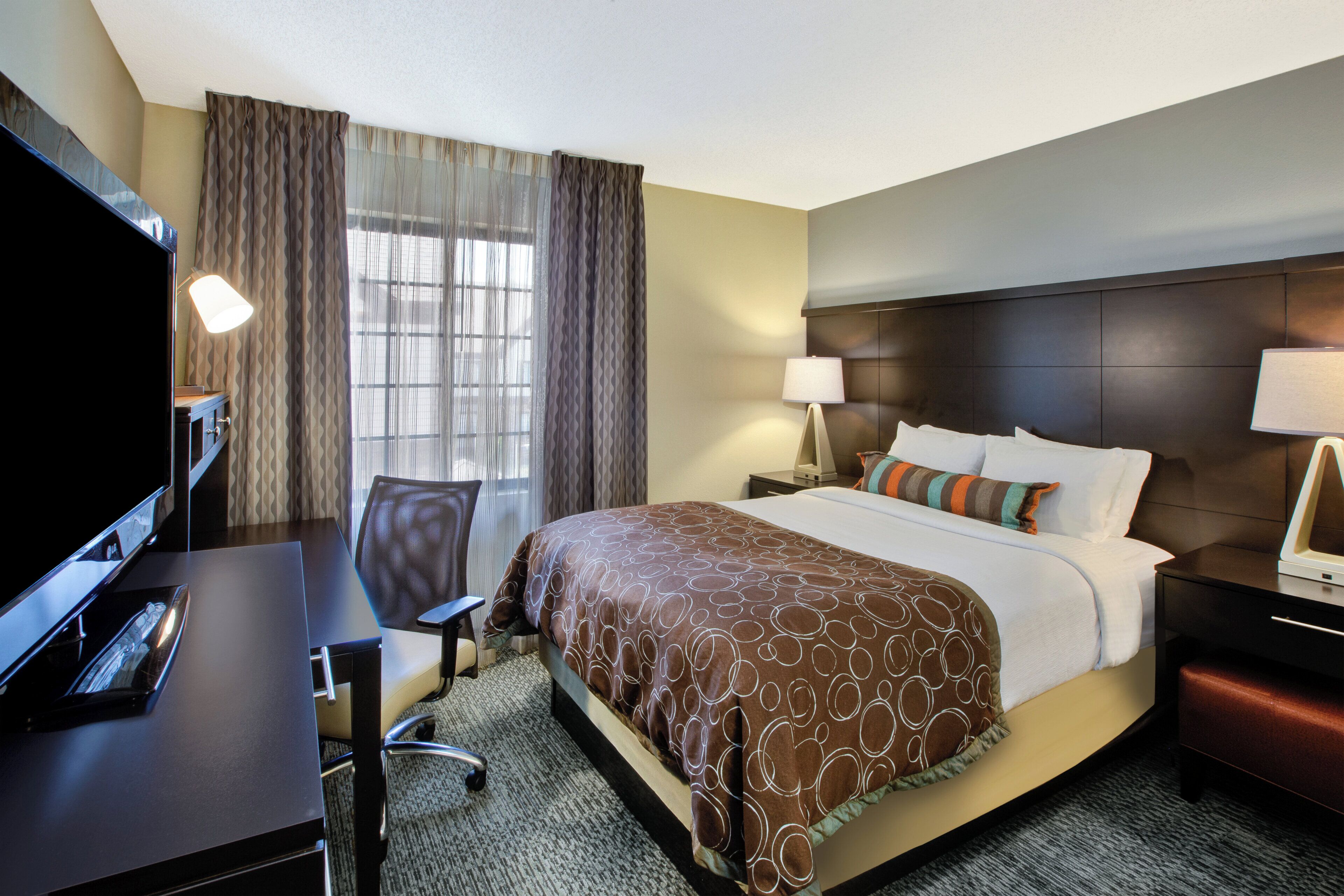 A hotel room with a bed, a desk, a chair, a TV, and a window with curtains.