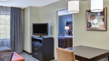 Una habitación de hotel con sofá, una mesita, televisor en mueble bajo, un cuadro en la pared y una ventana con cortinas.
