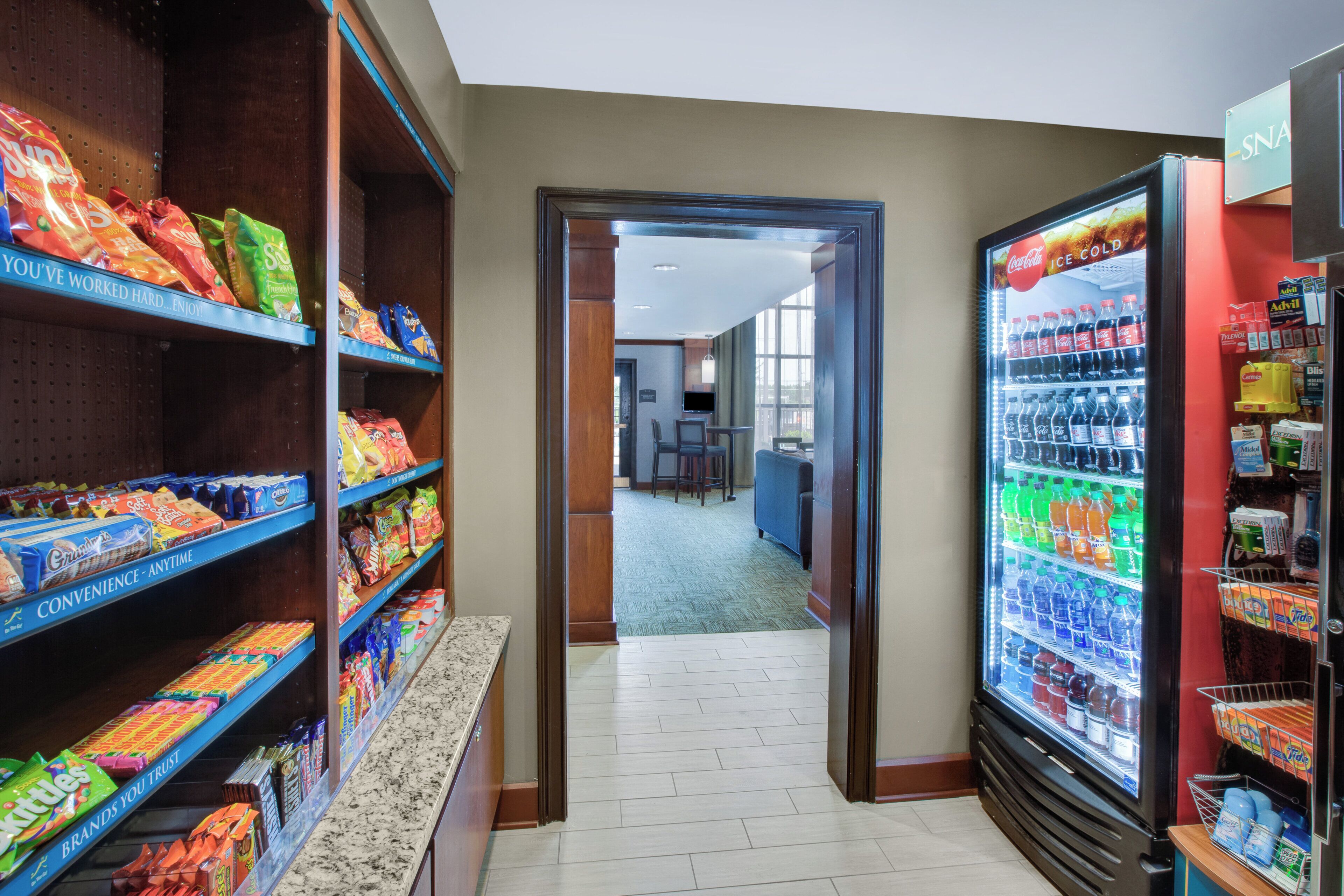 A corridor with snack shelves on the left, a refrigerator stocked with beverages on the right, and a glimpse of a dining area in the background.