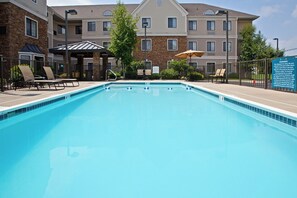 Une piscine rectangulaire bordée de carreaux, entourée de chaises longues et de parasols, avec un immeuble à plusieurs étages en arrière-plan.