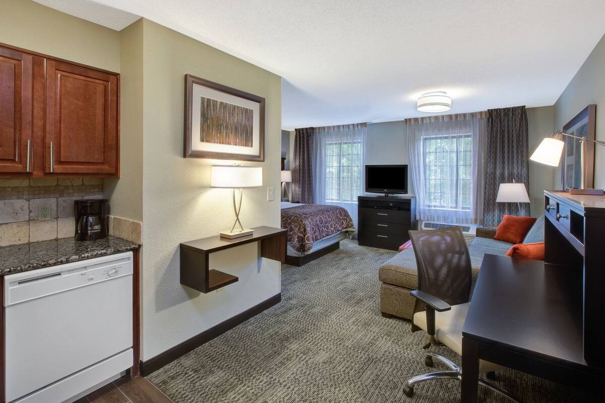 Une chambre d’hôtel équipée d’une kitchenette, d’un lit, d’un bureau, d’une chaise, d’une télévision et d’une fenêtre avec des rideaux.