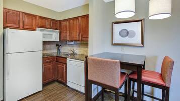 A compact kitchen with wooden cabinets, a white refrigerator, a microwave, and a dishwasher. There is a dining table with two chairs and a framed artwork on the wall.
