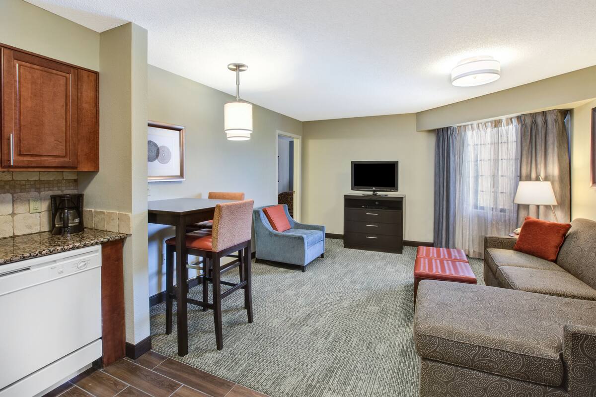 Une chambre d’hôtel compacte comprenant un coin cuisine, un espace repas, un coin salon avec un canapé et un fauteuil, un téléviseur à écran plat et une fenêtre avec des rideaux.