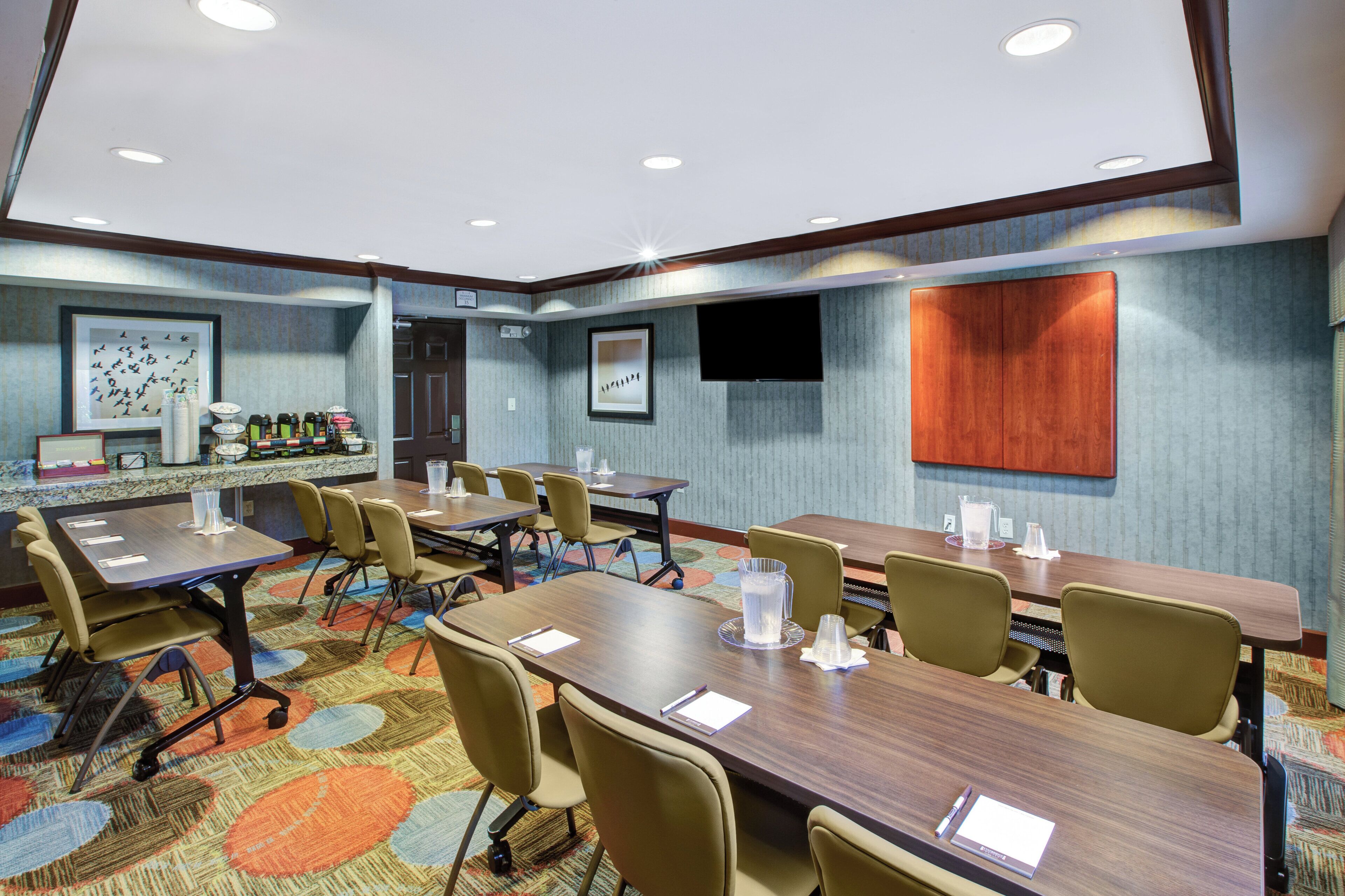 A conference room with a long table, multiple chairs, a coffee station, and a television on the wall.
