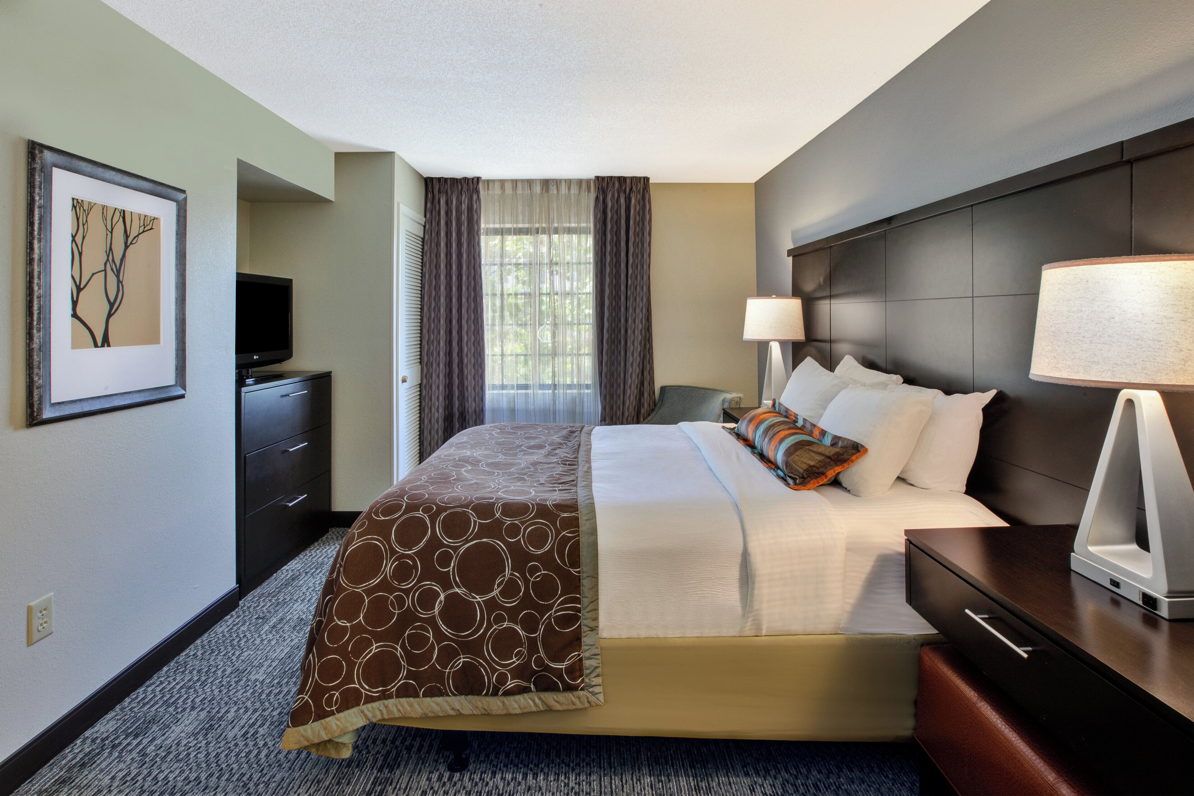 A hotel room with a bed, bedside tables, a TV, a window with curtains, and a painting on the wall.