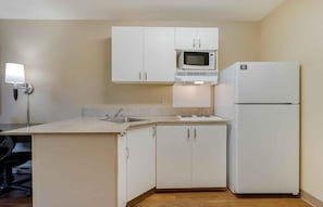 A small kitchenette with white cabinets, a microwave, a refrigerator, a sink, and a wall-mounted light.