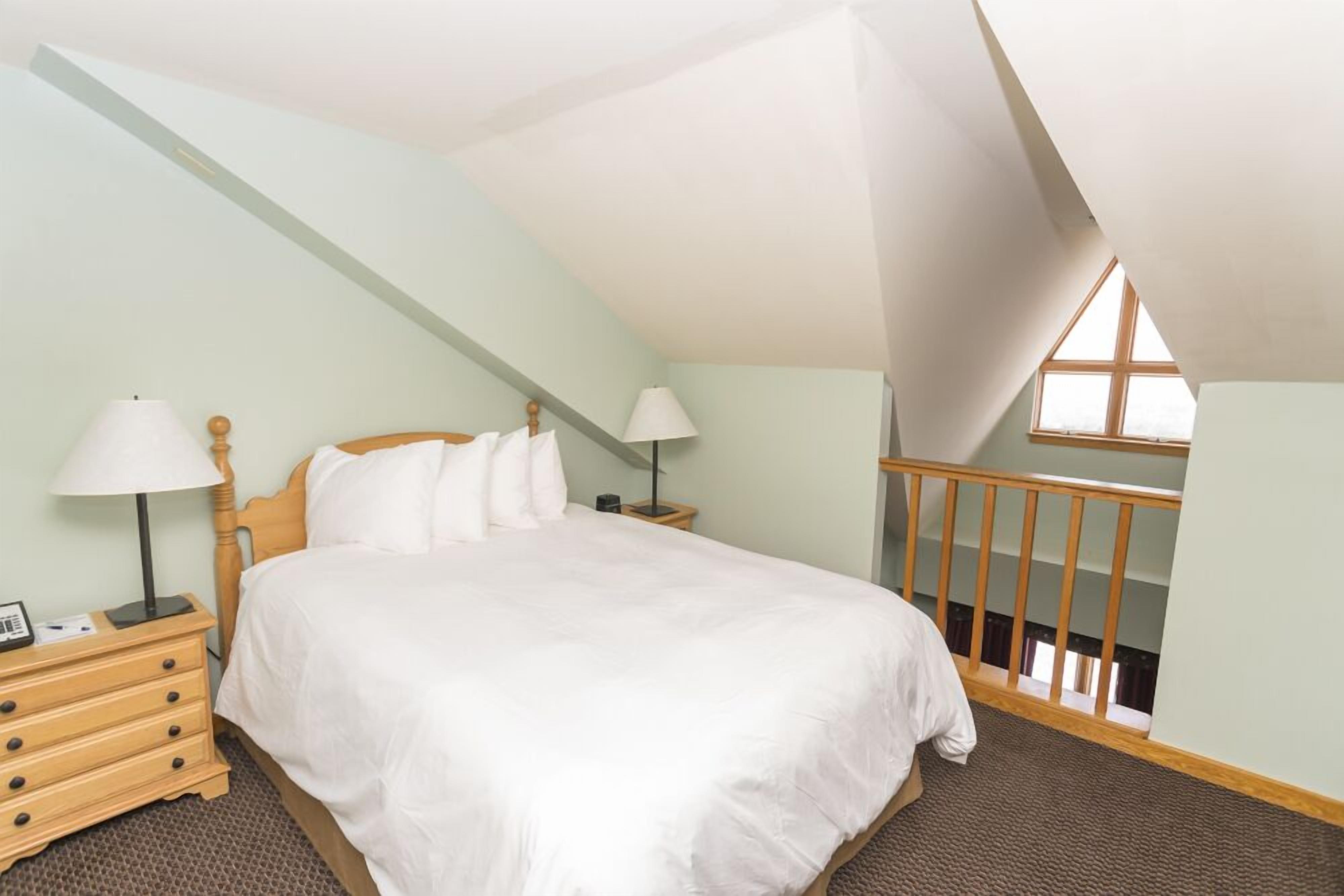 Une chambre avec un plafond en pente, un lit en bois, deux tables de chevet avec des lampes, une commode et une fenêtre donnant sur un beau paysage.