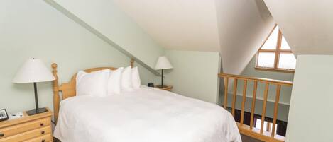 Une chambre avec un plafond en pente, un lit en bois, deux tables de chevet avec des lampes, une commode et une fenêtre donnant sur un beau paysage.