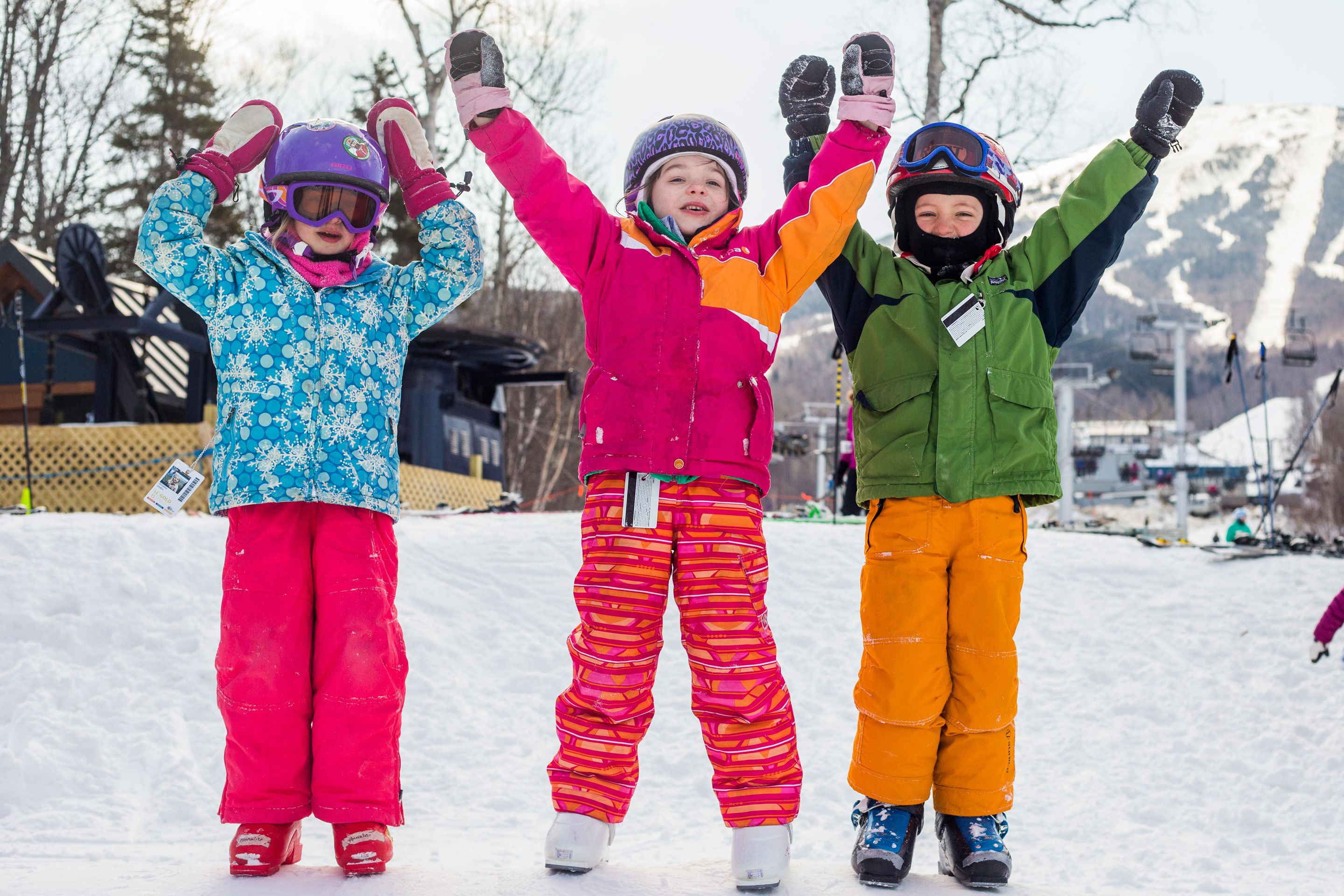 Three children in colorful winter clothing standing in a snowy area with a ski lift in the background.