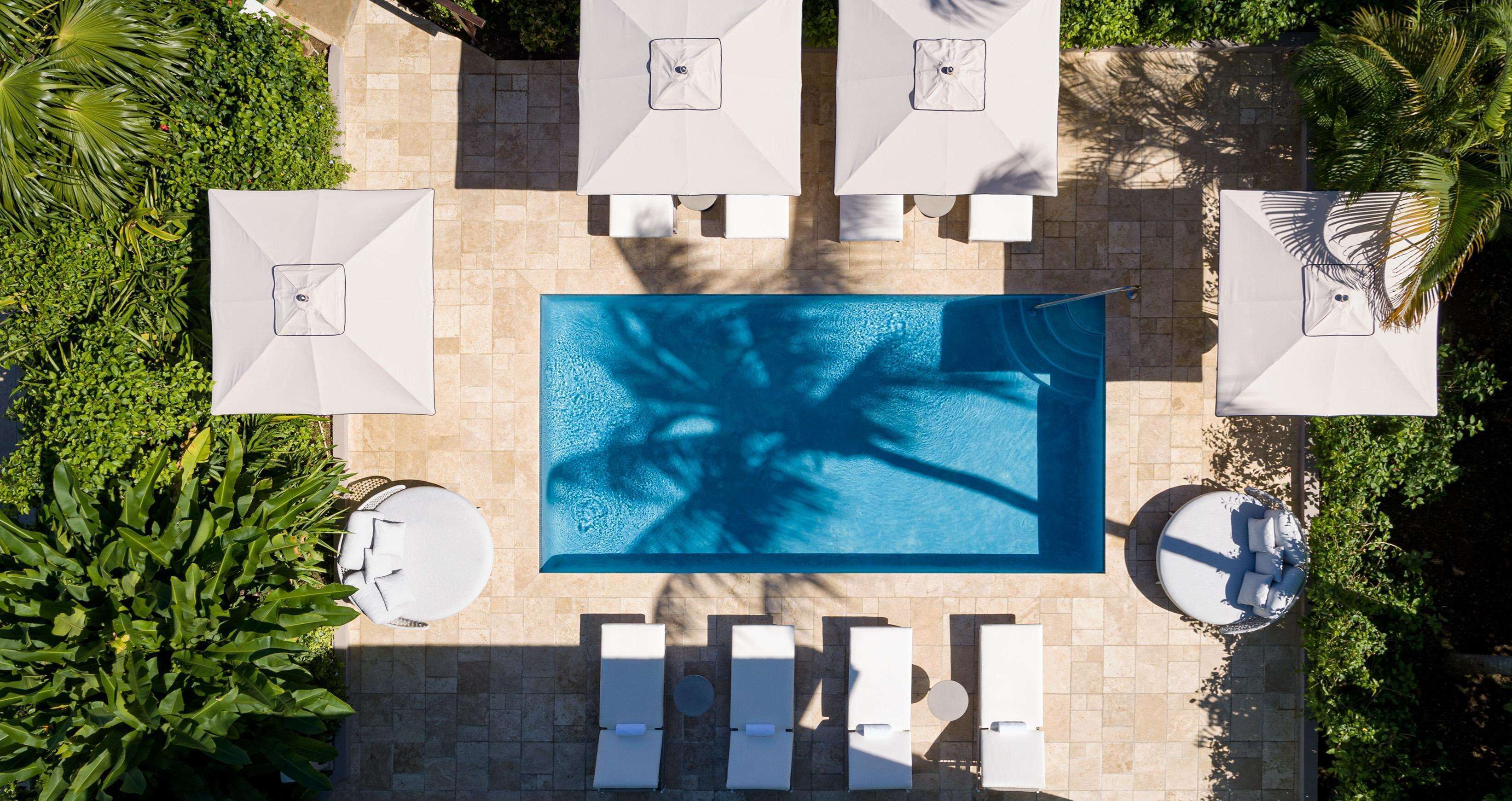 An aerial view of a rectangular swimming pool surrounded by white lounge chairs and umbrellas, with a tiled patio area and lush greenery.