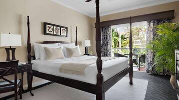 A four-poster bed with white linens, two bedside tables with lamps, a large window with curtains, and a view of greenery outside.