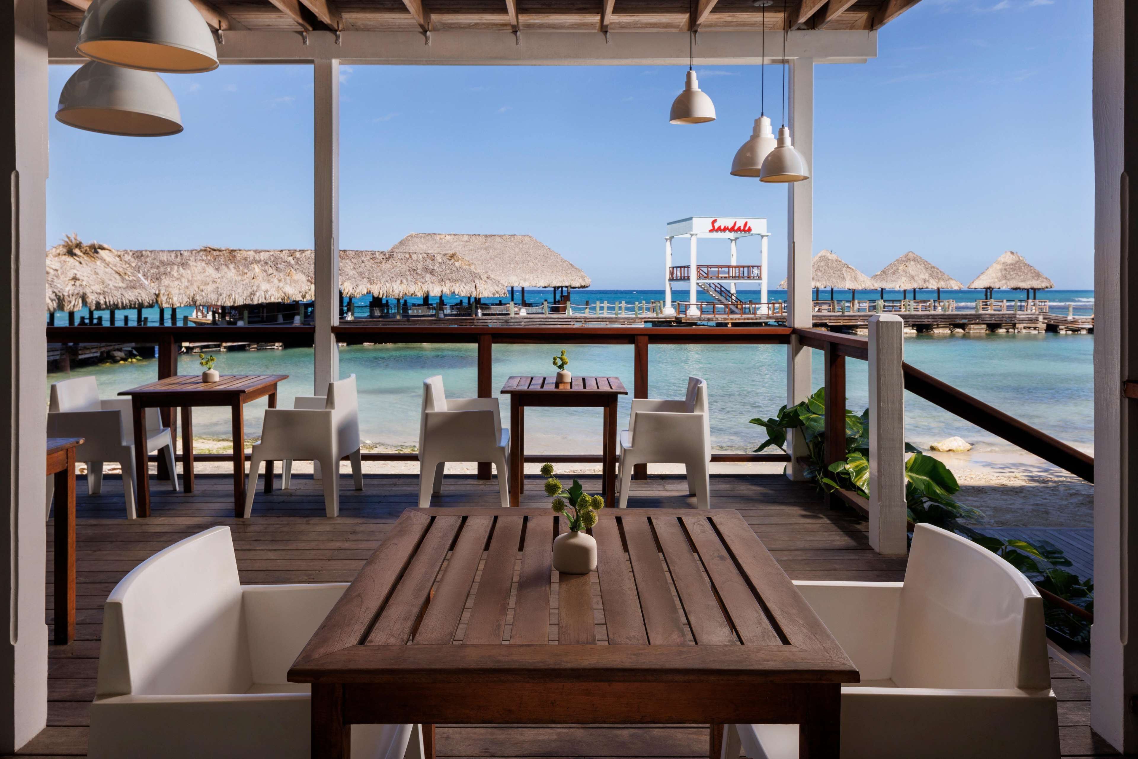 A wooden deck with white chairs and tables overlooking a beach with thatched umbrellas and a sign reading "Sundale".