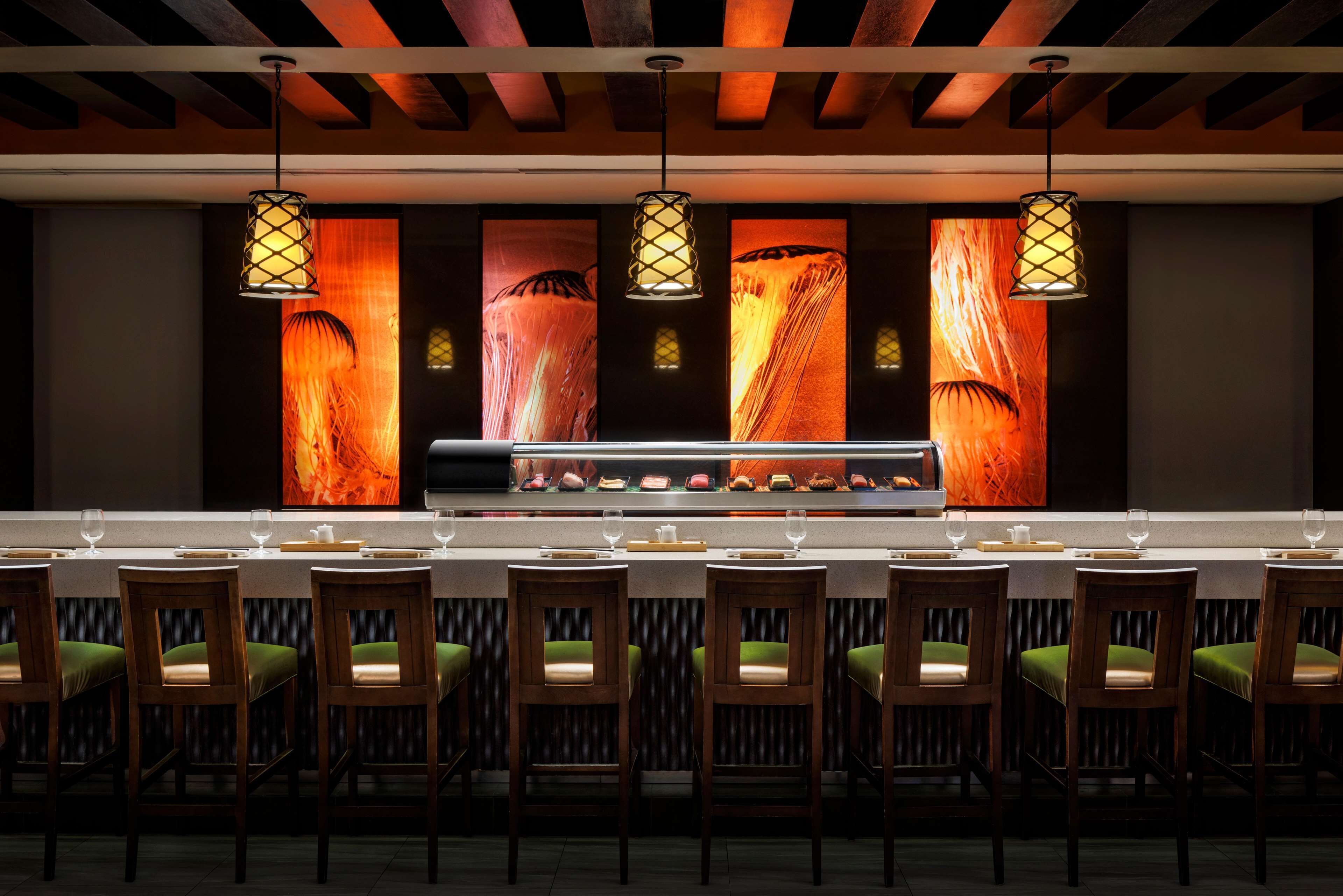 A restaurant with a long counter, wooden chairs with green cushions, and pendant lights with a patterned design.