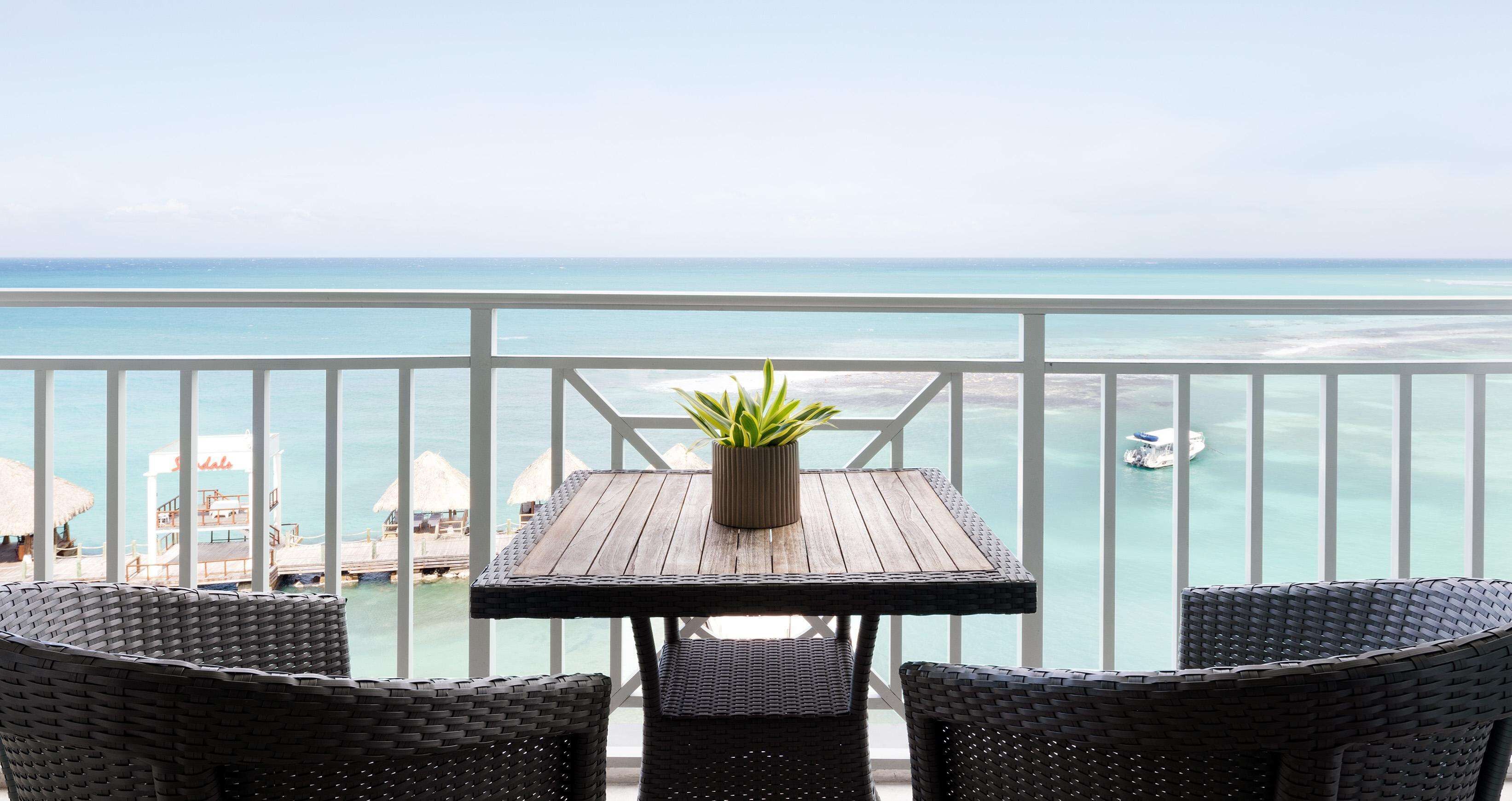 Ein Balkon mit Holztisch und Korbstühlen, mit Blick auf das türkisfarbene Meer und eine Hotelanlage.