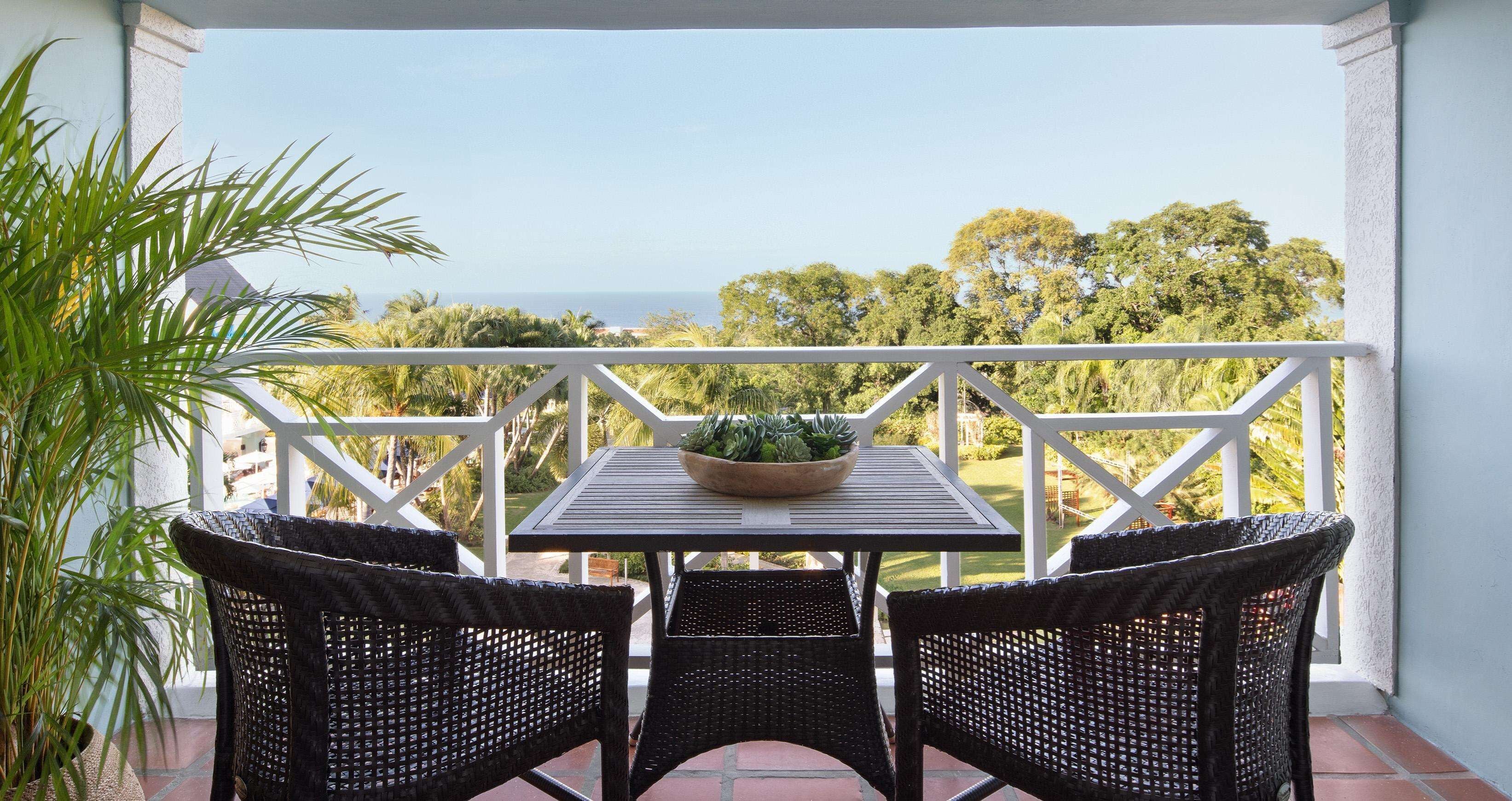 A balcony with a table and two wicker chairs, overlooking a lush landscape and the sea.