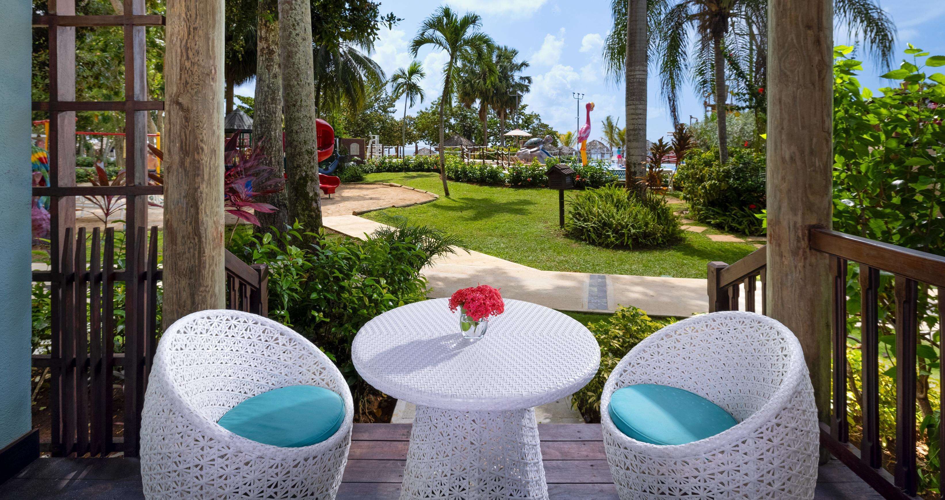 A covered outdoor seating area with wicker chairs and a small table, overlooking a landscaped garden with palm trees and a playground.