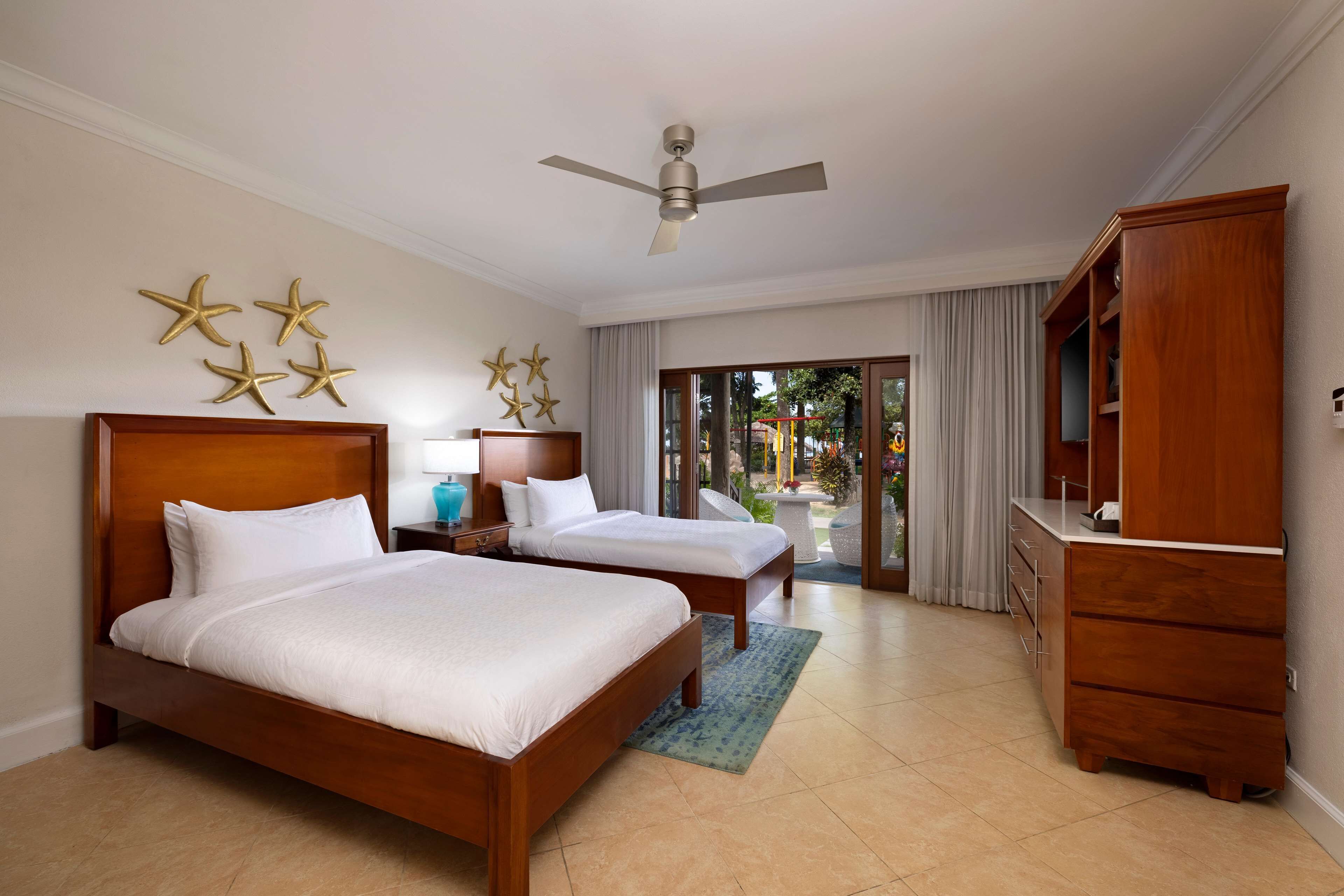 Habitación de hotel con dos camas, un ventilador de techo, un aparador de madera y vistas al exterior.