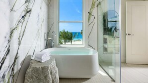 A modern bathroom with a large bathtub, a glass shower enclosure, and a view of the beach through a window.