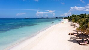 A beach with clear turquoise water, white sand, and palm trees. There are lounge chairs and umbrellas along the shore.