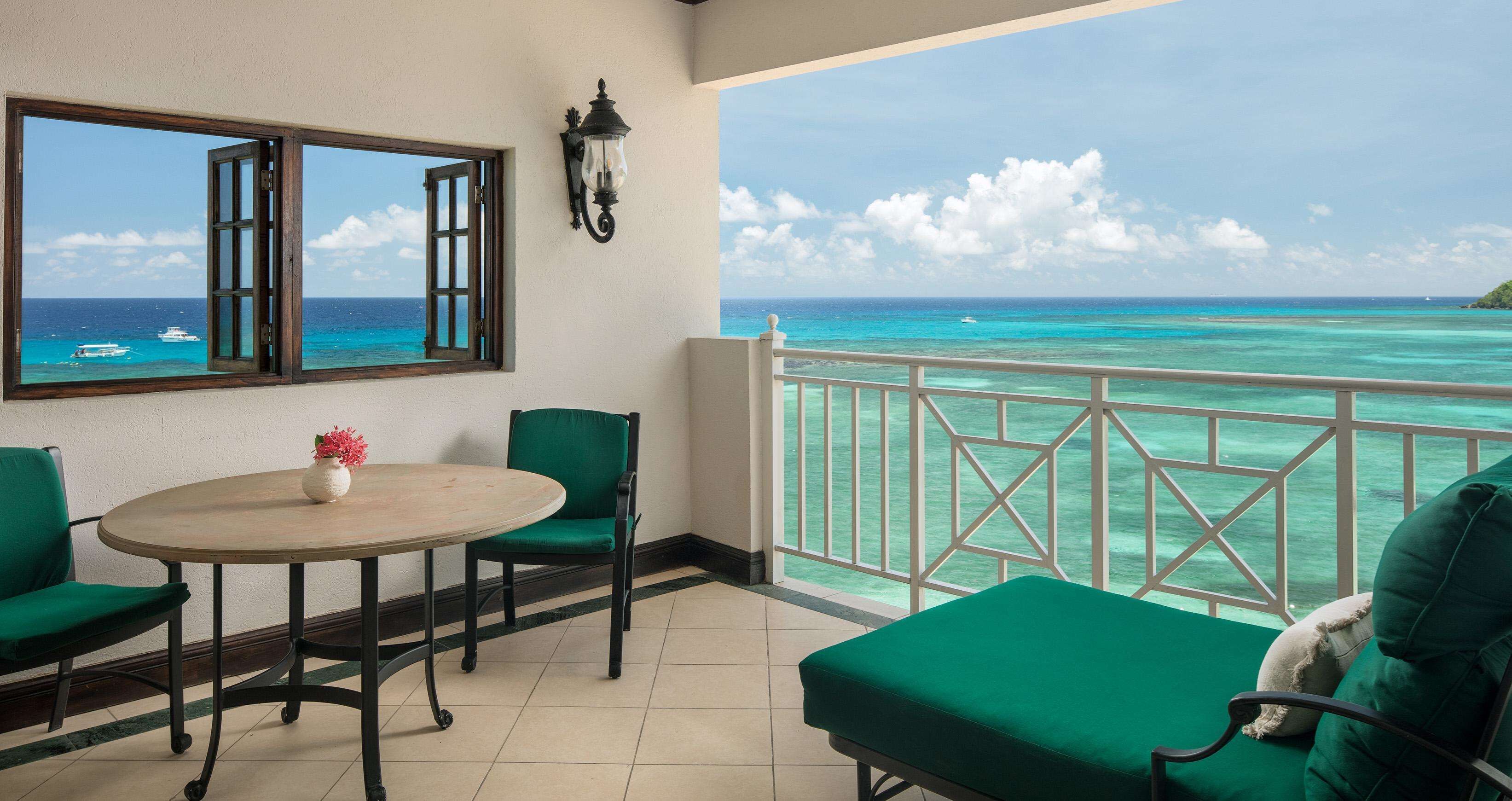 A balcony with a view of the ocean, a table with a vase, and green chairs.