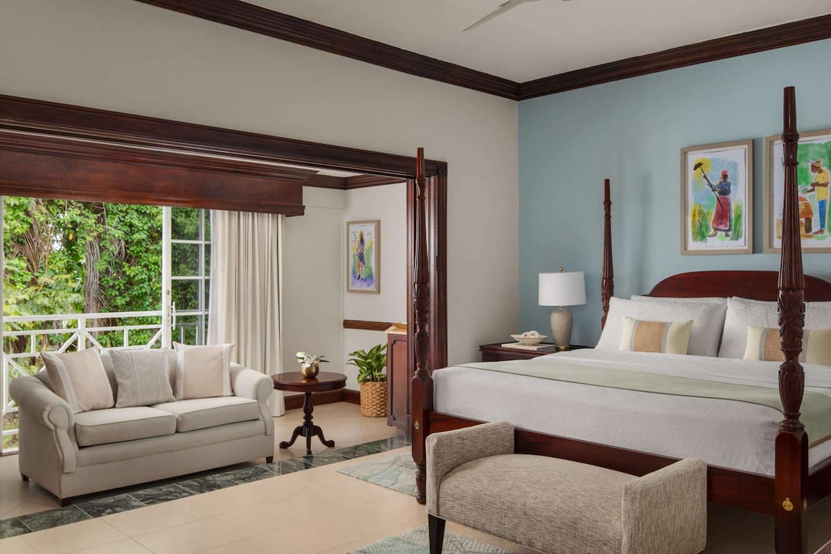 A spacious bedroom with a four-poster bed, a sitting area with a sofa, and a large window offering a view of greenery.