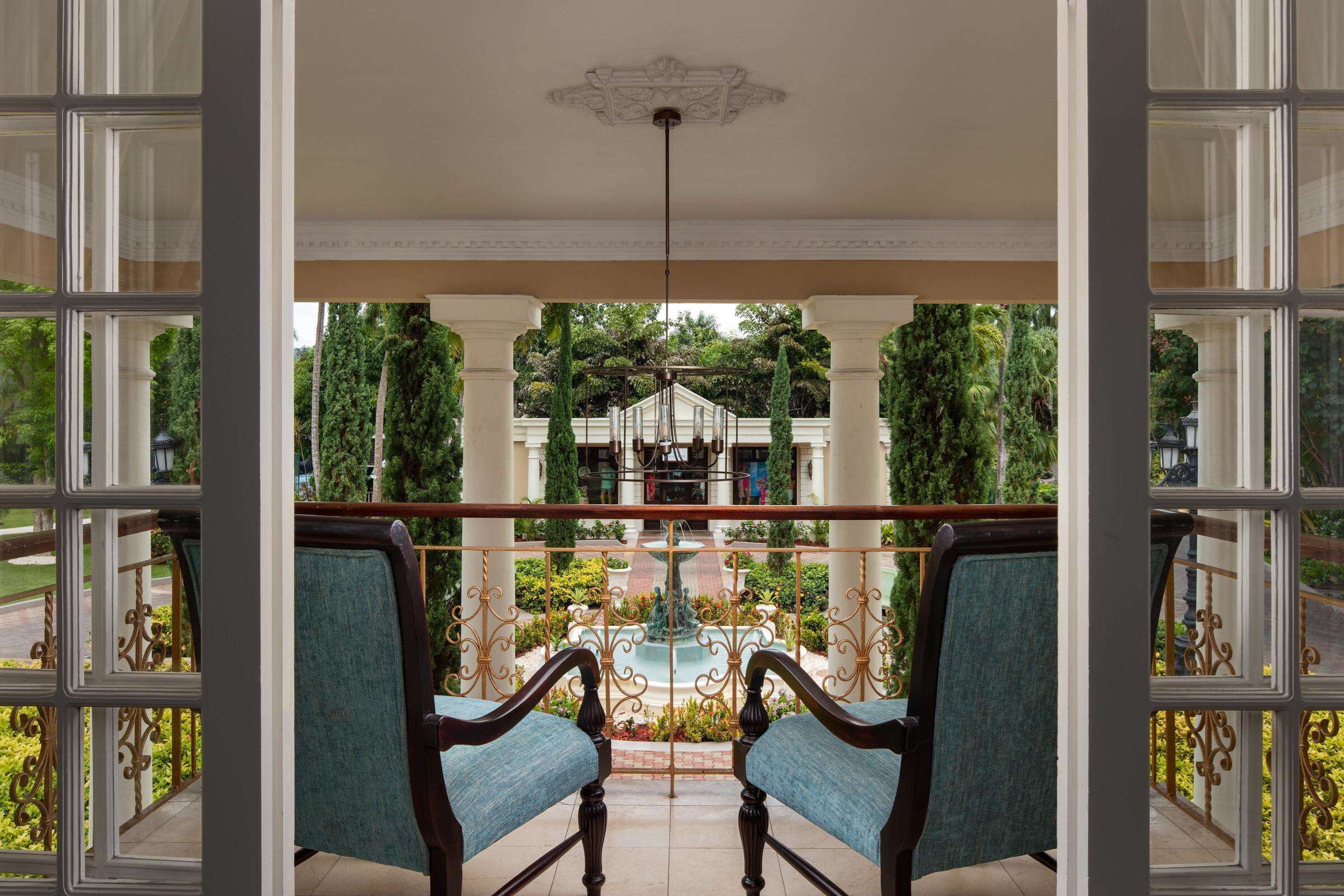 A balcony with two chairs, a fountain, and a view of a garden and building in the background.